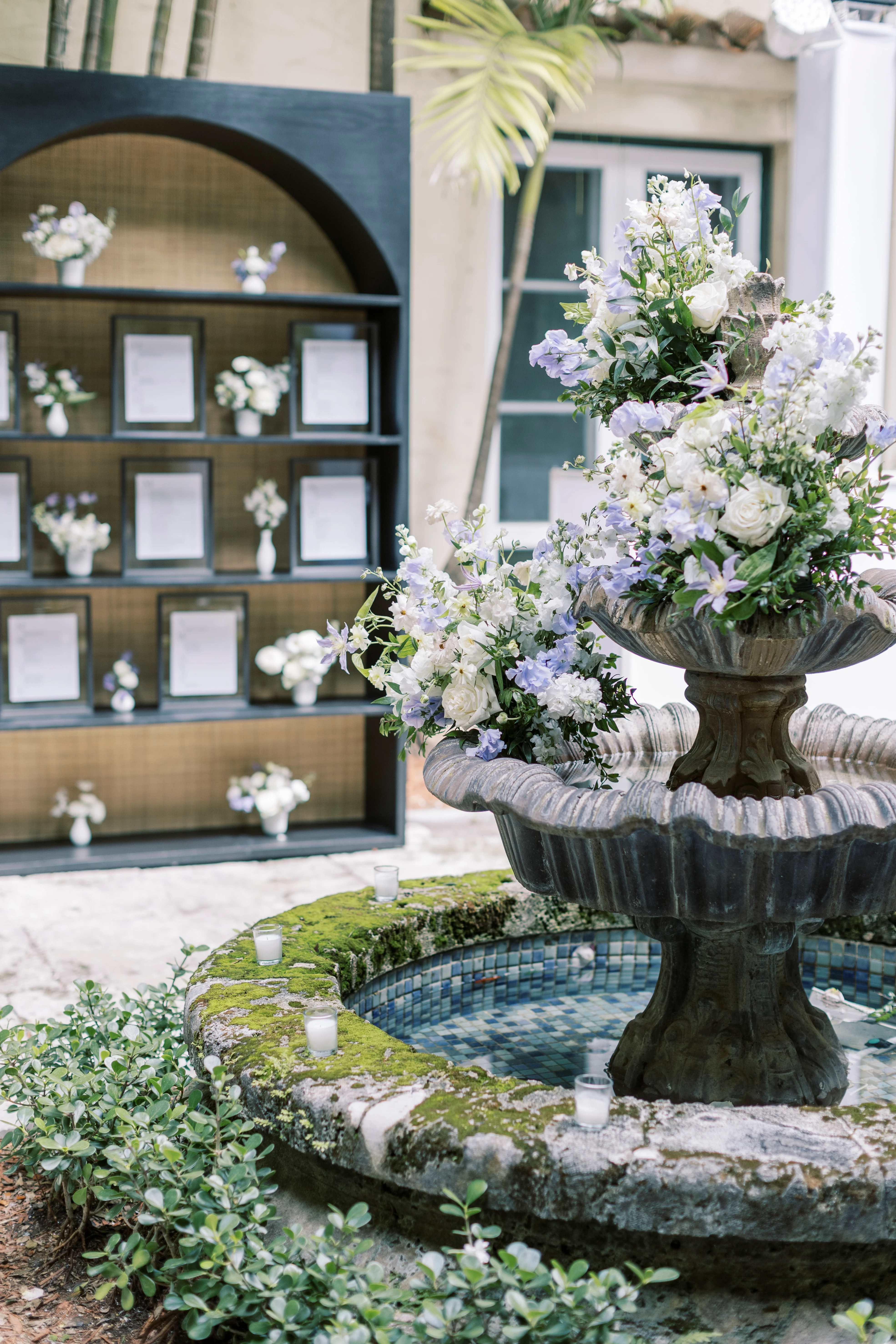 A bookshelf behind a water fountain with flowers and candles.